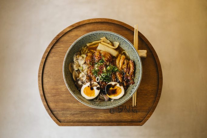 Bol de ramen avec œufs, légumes et viande sur une planche en bois.
